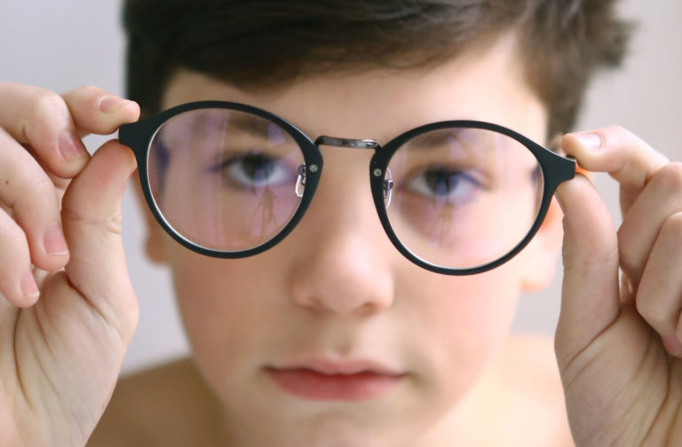 Close-up of a boy holding eyeglasses in front of his face, with his eyes visible through the lenses, representing vision correction or eye care for children.