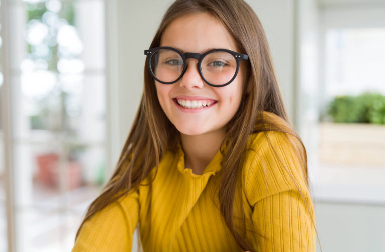 Smiling young girl wearing large round glasses and a yellow sweater, representing confidence and healthy vision in children.