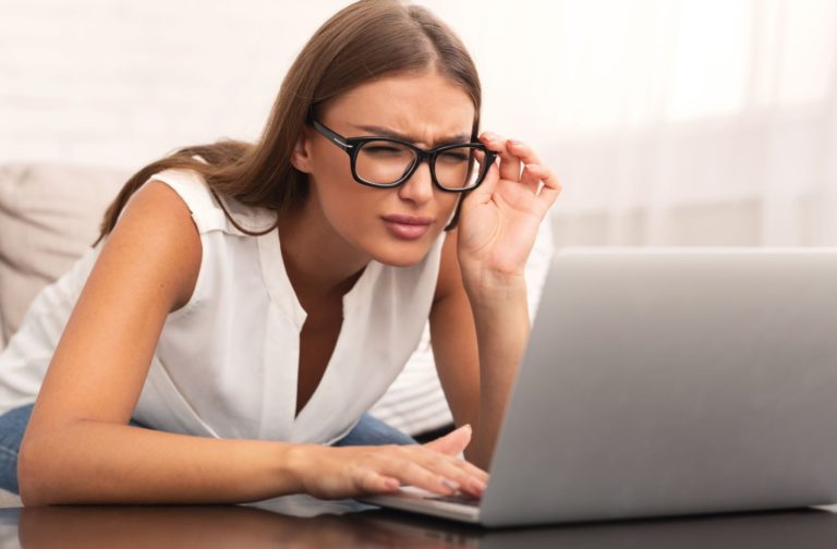 Young woman squinting and leaning in close to her laptop screen while adjusting her glasses, indicating difficulty seeing clearly from a distance, a common sign of myopia.