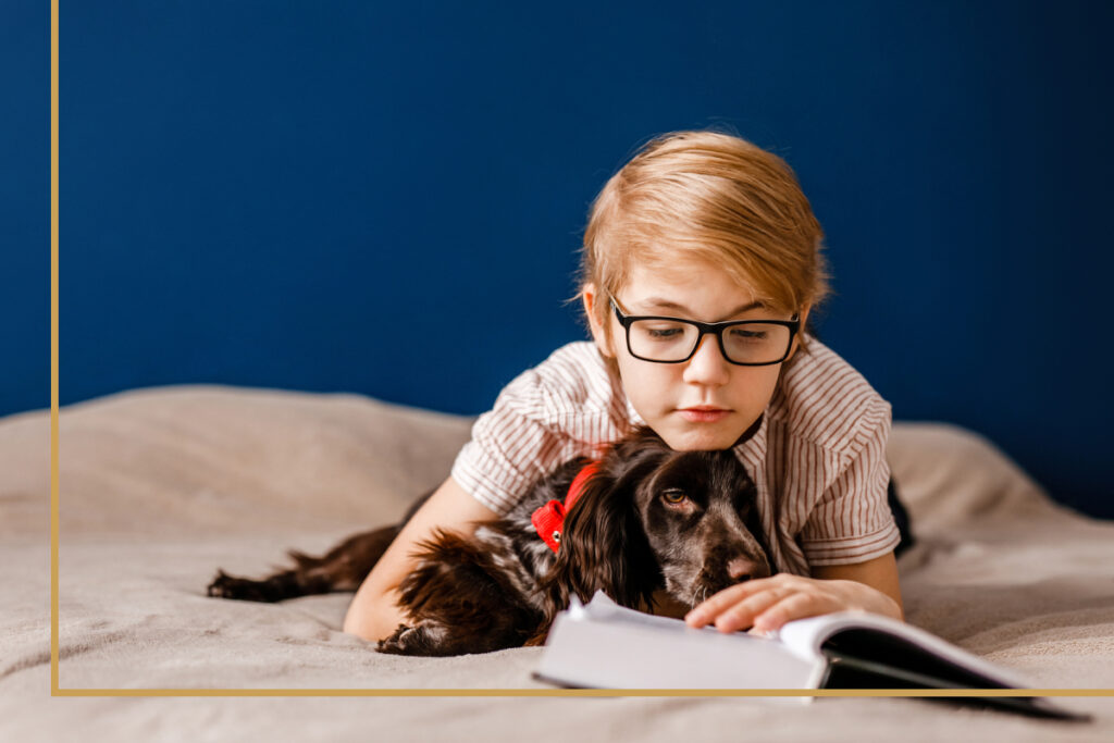 Young boy wearing glasses cuddling a small black and brown dog on a bed, in a cozy room with a blue wall — pediatric eyewear from Eyes Harvest Hills.